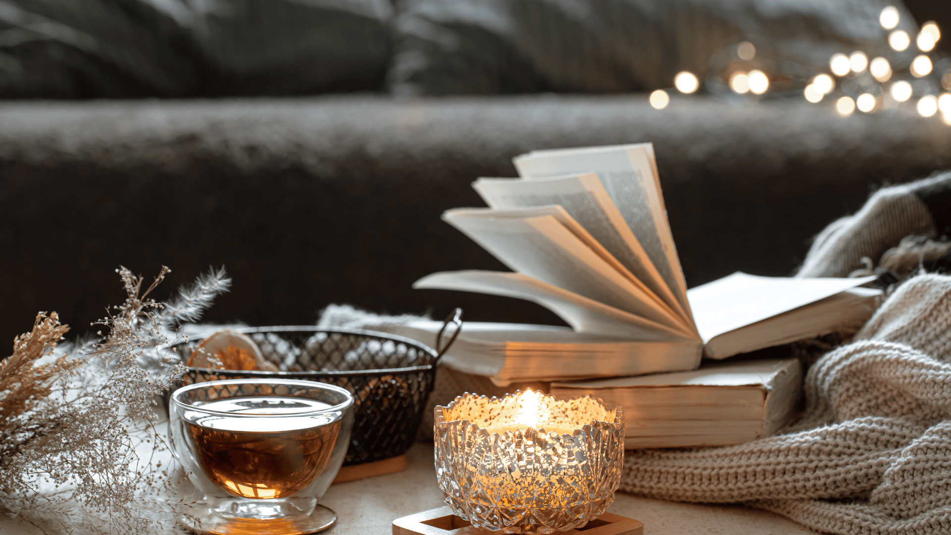 Gemütliche Szene mit einem offenen Buch, einer brennenden Kerze in einem verzierten Glasbehälter, einer Tasse Tee und weichen Textilien im Hintergrund, stimmungsvolle Beleuchtung mit warmen Lichtakzenten.
