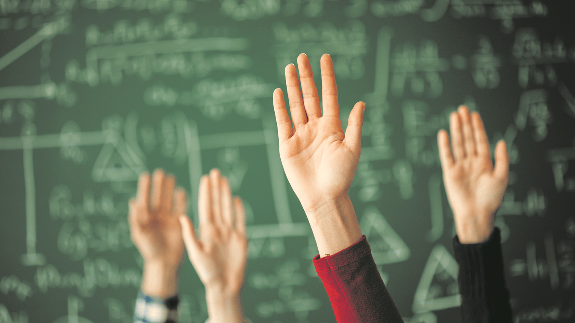 Mehrere Hände, die in einem Klassenzimmer vor einer Tafel mit mathematischen Formeln in die Luft gehoben sind