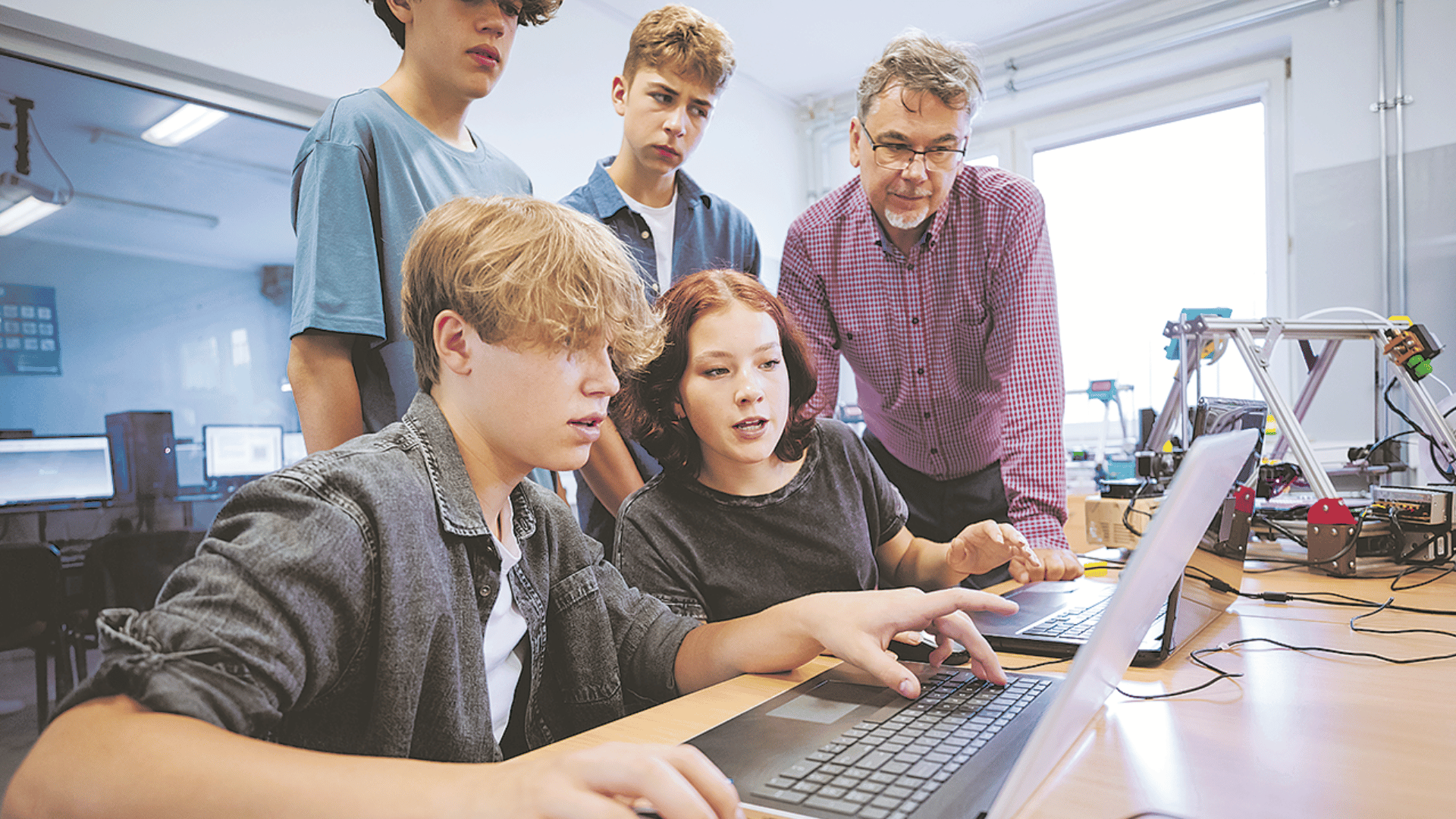 Eine Gruppe von Schülern und ein Lehrer in einem Klassenzimmer mit Computern. Die Schüler arbeiten gemeinsam an Laptops, während der Lehrer sie unterstützt.