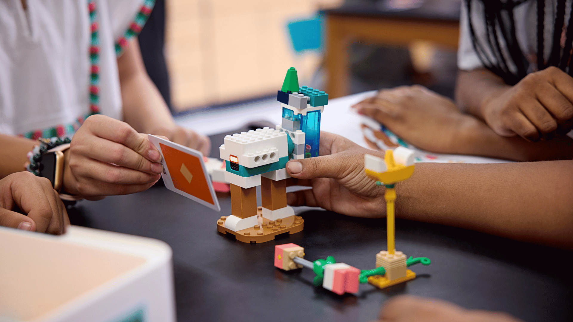 Kinderhände bauen und spielen mit LEGO Education SPIKE-Elementen auf einem Tisch. Sie halten eine Karte und konstruieren kreative Modelle aus bunten LEGO-Steinen.