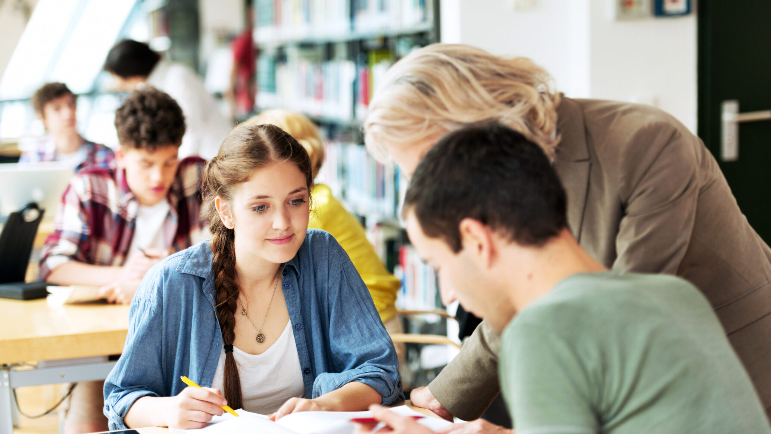 Schüler und Schülerin sitzen an einem Tisch im Klassenzimmer, während ihnen die Lehrerin etwas erklärt