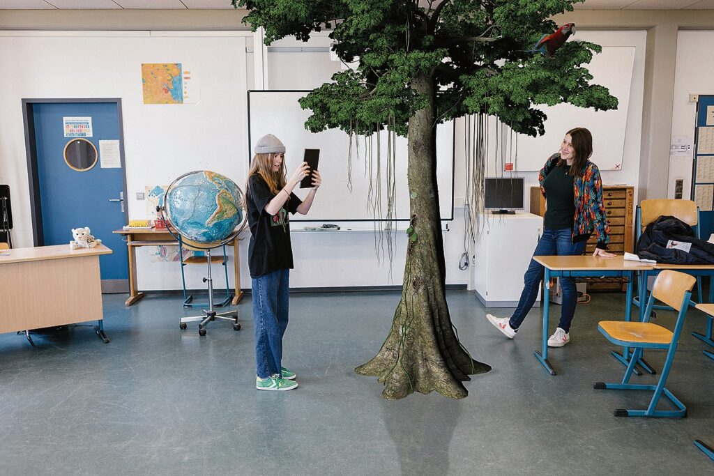 Schülerin steht vor einem Baum, der im Klassenzimmer wächst und betrachtet ihn durch ein Tablet